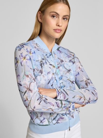 Modelka kobieca nosi Strickjacke mit Zweiwege-Reißverschluss von Marc Cain z mieszanki bawełny, prosty krój, prążkowany kołnierz, jasnoniebieskie i fioletowe kwiaty, prążkowane wykończenia, długość tyłu 55 cm, długość rękawa 78 cm w rozmiarze 36, zdjęcie: zbliżenie górnej części ciała z przodu