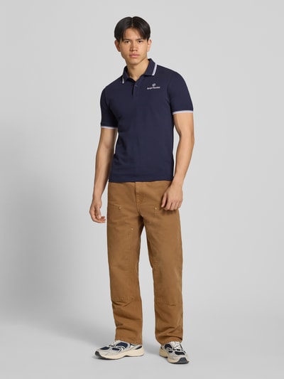 Męski model nosi Slim Fit Poloshirt mit Logo-Stitching Modell 'Reed' od SERGIO TACCHINI, elastyczna mieszanka bawełny, krótkie rękawy, polowy kołnierzyk, krój slim fit, granatowy kolor, przedni widok całej sylwetki.