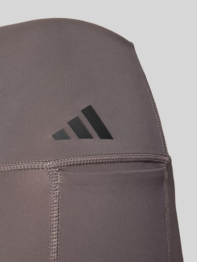 Vrouwenmodel draagt Leggings met Label-Print van adidas Sportswear, taupe kleur, stretchstof, elastische tailleband, detailopname heup.