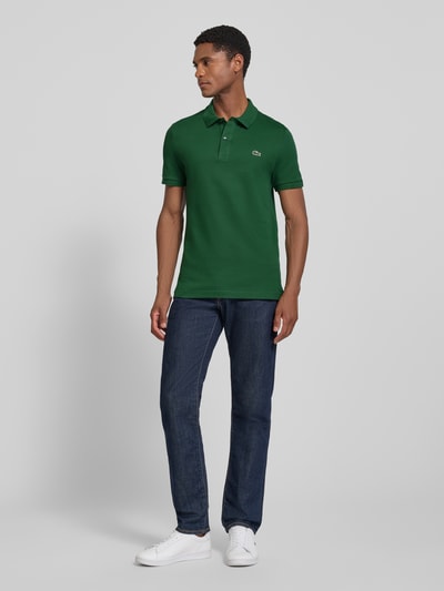 Männermodel trägt Slim Fit Poloshirt aus reiner Baumwolle von Lacoste in Dunkelgrün mit glatter Struktur, Ganzkörperansicht von vorne.