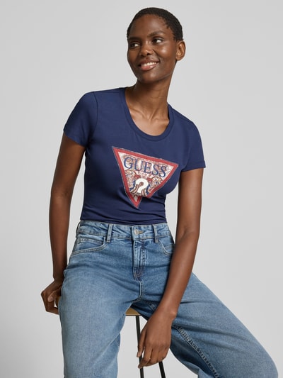 Vrouwenmodel draagt T-shirt met logo en ronde hals van guess, mix van katoen en elastaan, slim fit, logoprint, ruglengte maat s 58 cm, close-up bovenlichaam.