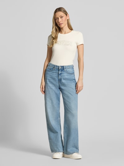 Frauenmodel trägt Slim Fit T-Shirt mit Strasssteinbesatz von Guess, Cremefarbe, Baumwoll-Mix, Logo-Detail, Rundhalsausschnitt, Ganzkörperansicht von vorne.