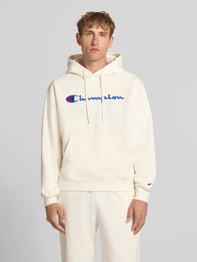 Männermodel trägt Hoodie mit Label-Stitching von Champion, cremefarben, Baumwoll-Mix, lockerer Schnitt, Ganzkörperansicht von vorne.