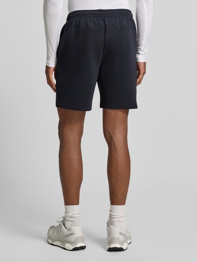 Męski model nosi szorty z kieszenią zapinaną na zamek błyskawiczny od Under Armour, mieszanka bawełny, krój regular fit, logo, elastyczny pas, szerokość w rozmiarze M: 39 cm, długość wewnętrzna nogawki w rozmiarze M: 18 cm, ciemnoniebieski kolor, gładka tekstura, całkowity widok sylwetki z tyłu.