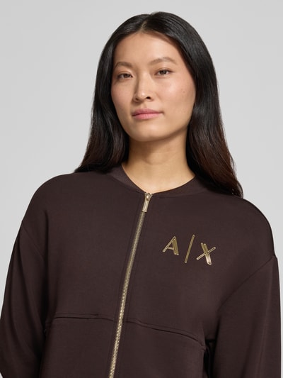 Frauenmodel trägt Sweatjacke mit Label-Print von Armani Exchange in Dunkelbraun mit goldenem Zweiwege-Reißverschluss, Nahaufnahme Oberkörper, Viskose-Mix mit Stretch-Anteil, lockere Passform, seitliche Eingrifftaschen.