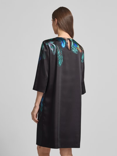 Frauenmodel trägt Relaxed Fit Kleid mit Foto-Print von Marc Cain in Schwarz mit bunten Federmotiven, elastischer Material-Mix, Ganzkörperansicht Rückansicht.