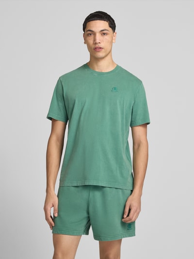 Męski model nosi T-Shirt mit Label-Stitching und Rundhalsausschnitt od CHAMPION z bawełny, krój regular fit, długość tyłu 67 cm, zielony kolor, gładka faktura, Ganzkörperansicht von vorne.