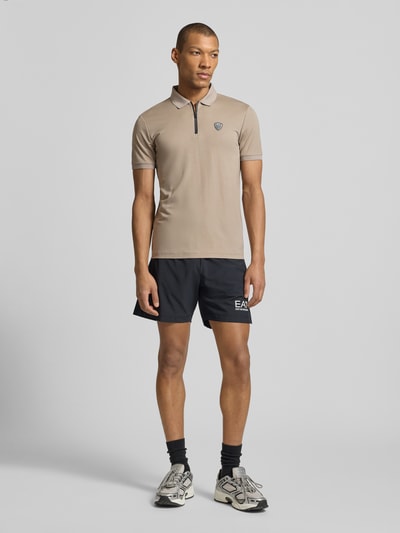 Męski model nosi Slim Fit Poloshirt aus Viskose-Mix von EA7 Emporio Armani, elastyczna mieszanka wiskozy, krój slim fit, detal z logo, kołnierzyk polo, elastyczne zakończenia rękawów, długość tyłu w rozmiarze M: 60 cm, całopostaciowy widok z przodu