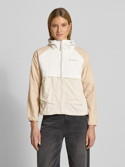 Frauenmodel trägt Windbreaker mit Rückenbelüftungssystem und Kapuze von COLUMBIA in Beige und Weiß, Lockerer-Schnitt, Omni-Shield™, Wasserabweisend, Kinnschutz, Eingrifftaschen mit Reißverschluss, Elastische Bündchen, Saum mit Kordelzug, Logo-Print; Ganzkörperansicht von vorne.