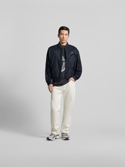 Männermodel trägt Relaxed Fit Jacke mit Logo-Print von MM6 Maison Margiela in Dunkelblau, elastische Abschlüsse, Reißverschluss, Ganzkörperansicht von vorne.