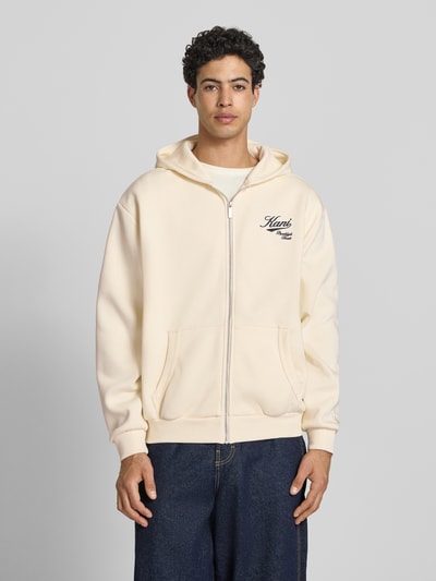 Männermodel trägt Oversized Sweatjacke mit Logo-Stitching von KARL KANI aus Baumwoll-Mix, Kapuze, Kängurutasche, Reißverschluss, cremefarben, Oversized-Fit, elastische Abschlüsse, Rückenlänge 62 cm, Ärmellänge 65 cm, Ganzkörperansicht von vorne.