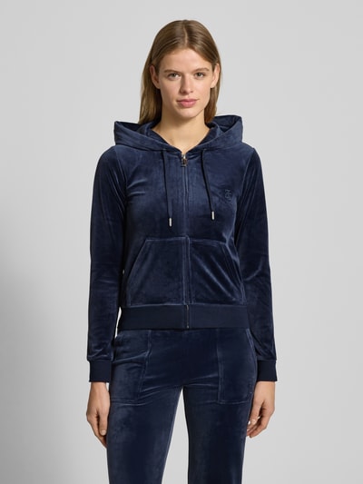 Frauenmodel trägt Sweatjacke mit Label-Stitching Modell 'ROBERTSON' von Juicy Couture aus Material-Mix mit Stretch-Anteil, Regular-Fit, Kapuze mit Tunnelzug, Reißverschluss und Känguru-Tasche in Dunkelblau, samtige Textur, Ganzkörperansicht von vorne.