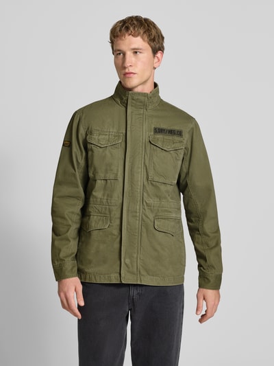 Männermodel trägt Military-Jacket mit Pattentaschen und Label-Patches von Superdry in Oliv, Gerader-Schnitt, Stehkragen, Ganzkörperansicht von vorne. Rückenlänge bei Größe M: 72 cm, Schulterbreite: 46 cm, Brustweite: 59 cm, Ärmellänge: 66 cm, Klettverschluss, Reißverschluss, Label-Patches.