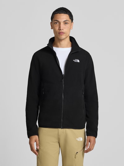 Męski model nosi czarną kurtkę z polaru z zamkiem błyskawicznym, Model 'GLACI' od The North Face, taliowany krój, kieszenie zapinane na zamek błyskawiczny, wyhaftowane logo, długość tyłu w rozmiarze M: 66 cm, długość rękawów w rozmiarze M: 68 cm, widok całej sylwetki od przodu.