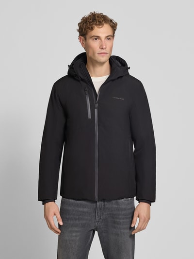 Męski model nosi Regular Fit kurtka z nadrukiem z logo 'Regular Fit Jacke mit Logo-Print' od Lindbergh, elastyczna mieszanka materiałów, kaptur z tunelem, napoleońska kieszeń, czarna, gładka faktura, długość tyłu w rozmiarze M: 74 cm, szerokość ramion: 46 cm, szerokość w rozmiarze M: 42 cm, długość rękawów: 62 cm, widok całej sylwetki z przodu.