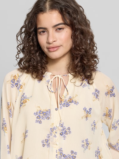 Frauenmodel trägt Relaxed Fit Chiffon-Bluse mit Allover-Muster Modell 'FUSINA' von Only mit Rundhals, Raffungen, Bindeband, Ballonärmel, Knopf mit Öse, Materialverarbeitung: Chiffon, zart cremefarben mit lila und gelben Blumenmotiven, Nahaufnahme Oberkörper.
