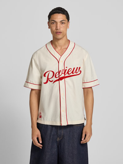 Männermodel trägt Relaxed Fit Freizeithemd mit Logo von REVIEW in Reine Baumwolle, V-Ausschnitt, Ganzkörperansicht von vorne, Rückenlänge 74 cm, Schulterbreite 50 cm, Ärmellänge 29 cm, Farben Creme mit roten Akzenten, glatte Textur.