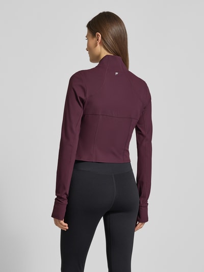 Frauenmodel trägt Slim Fit Jacke mit stójka model 'Storm' od Fabletics, krój krótki, pętelki na kciuki, kolor bordowy, Ganzkörperansicht Rückansicht. Długość tyłu w rozmiarze S: 44 cm, szerokość ramion w rozmiarze S: 36 cm, szerokość klatki piersiowej w rozmiarze S: 42 cm, długość rękawów w rozmiarze S: 74 cm.
