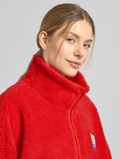 Frauenmodel trägt Oversized Teddyjacke mit Stehkragen Modell 'HOKTOWN' von American Vintage, leuchtend rot, weiche Teddy-Textur, Nahaufnahme Oberkörper, Damen Teddyjacke, Reißverschluss, Label-Detail, Eingrifftaschen, Kontext: Rückenlänge 89 cm, Schulterbreite 58 cm, Brustweite 67 cm, Ärmellänge 49 cm.