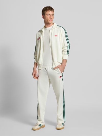 Männermodel trägt Sweatpants mit Reißverschlusstaschen und Logo-Stitching von adidas Originals, weiß, grünes Streifendetail, Material-Mix, lockerer Schnitt, elastischer Bund, Ganzkörperansicht von vorne.
