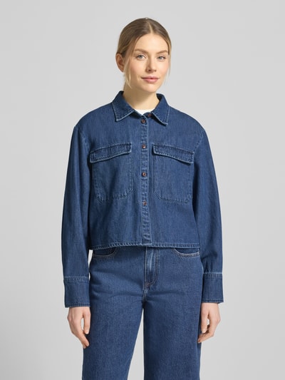 Vrouwenmodel draagt Cropped Jeansbluse aus reiner Baumwolle Modell 'POSEY' van Vero Moda in denim look, boxy fit, hemdblousekraag, lange mouw met manchet, borstklepzakken, eenrijige knoopsluiting, materiaal zuivere katoen. Vooraanzicht bovenlichaam.