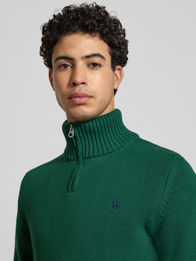 Männermodel trägt Slim Fit Pullover in Strick-Optik von Superdry in Grün mit Stehkragen, Reißverschluss, Logo-Stitching, Nahaufnahme Oberkörper.