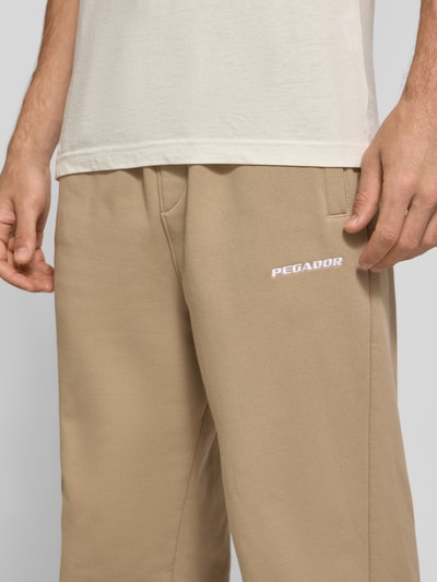 Männermodel trägt Loose Fit Sweatpants aus Baumwoll-Mix mit Label-Detail von PEGADOR in Beige, sichtbare Eingrifftaschen, Nahaufnahme Oberkörper. Herren Sweatpants, Elastikbund, Innenbeinlänge bei Größe M: 70 cm.