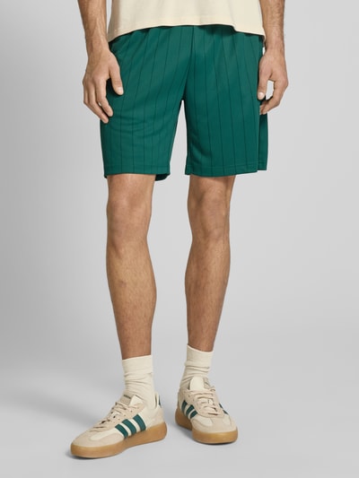 Männermodel trägt KARL KANI Shorts mit elastischem Bund Modell 'Insignia Shadow' im Allover-Look, Regular-Fit in Grün, Nahaufnahme Oberschenkel, Bundweite 38 cm, Innenbeinlänge 19 cm, Größe M.