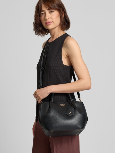 Frauenmodel trägt Handtasche in Leder-Optik Modell 'Amorette' von GUESS in Schwarz mit Logo-Detail, abnehmbarer Trageriemen, Ganzkörperansicht von vorne.
