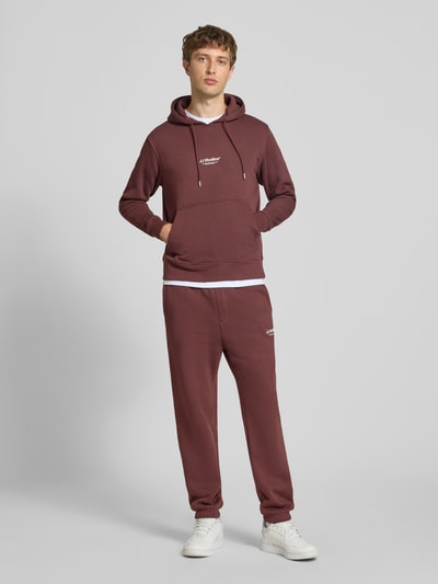 Männermodel trägt Regular Fit Hoodie aus Baumwoll-Mix Modell 'Soho' von JACK & JONES in Dunkelrot, Känguru-Tasche, Ganzkörperansicht von vorne. Rückenlänge 66 cm, Ärmellänge 65 cm, Kapuze mit Tunnelzug, elastische Abschlüsse.