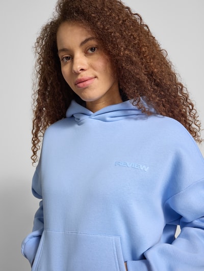 Frauenmodel trägt Hoodie mit Logo und Känguru-Tasche von Review, hellblau, Baumwoll-Mix, Relaxed Fit, Nahaufnahme Oberkörper.