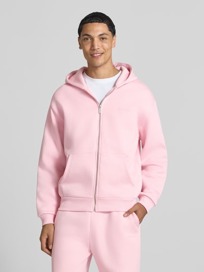 Männermodel trägt Sweatjacke aus Baumwoll-Mix von REVIEW in zartem Rosa, lockere Passform, Kängurutasche, Ganzkörperansicht von vorne.