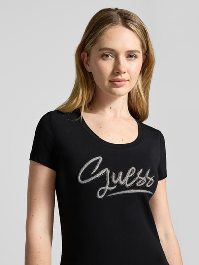 Vrouwenmodel draagt Slim Fit T-Shirt met Strasssteinbesatz van Guess in zwart, elastische katoenmix, logo-detail, ronde hals, close-up bovenlichaam.