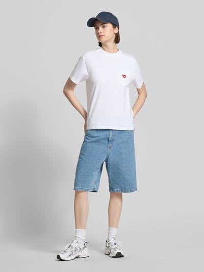 Frauenmodel trägt Damen T-Shirt mit Label-Patch von Carhartt Work In Progress aus Reine Baumwolle, Gerader-Schnitt, Rundhalsausschnitt, Brusttasche, Rückenlänge 58 cm bei Größe S, Farbkontrast durch weiße Töne und blaues Denim, glatte und strukturierte Stoffe, Ganzkörperansicht von vorne.