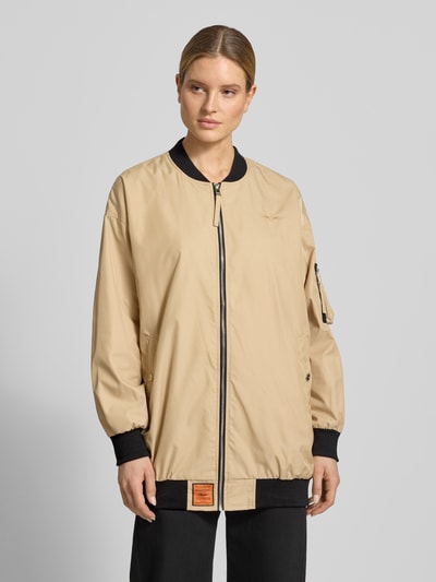 Frauenmodel trägt Relaxed Fit Jacke mit Logo-Stitching von Original Bombers, beige mit schwarzen Bündchen, glatte Textur, Ganzkörperansicht von vorne, Stehkragen, Reißverschluss, Armtasche, seitliche Eingrifftaschen mit Druckknopf, elastische Abschlüsse, Rückenlänge 84 cm, Schulterbreite 58 cm, Brustweite 56 cm, Ärmellänge 57 cm.