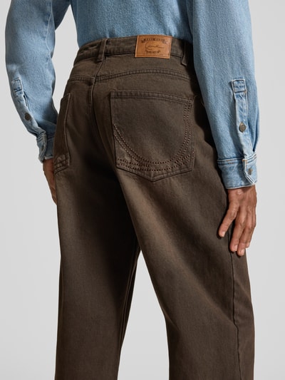 Männermodel trägt Relaxed Fit Jeans mit Knopf- und Reißverschluss von KARL KANI in Braun, 5-Pocket-Design, Label-Patch, Ganzkörperansicht Rückansicht.