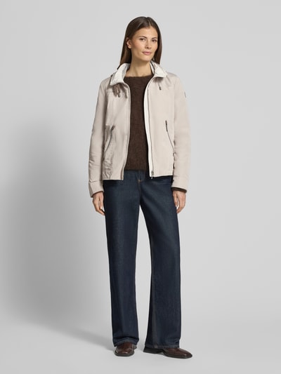 Modelka kobieca nosi Jacke mit Kapuze und Reißverschlusstaschen od Gil Bret, krój regular fit, beżowy kolor, kaptur z troczkiem, stójka, zamek błyskawiczny, kieszenie na zamek, długość tyłu w rozmiarze 36: 59 cm, szerokość ramion w rozmiarze 36: 40 cm, szerokość w biuście w rozmiarze 36: 51 cm, długość rękawa w rozmiarze 36: 63 cm, pełny widok z przodu.