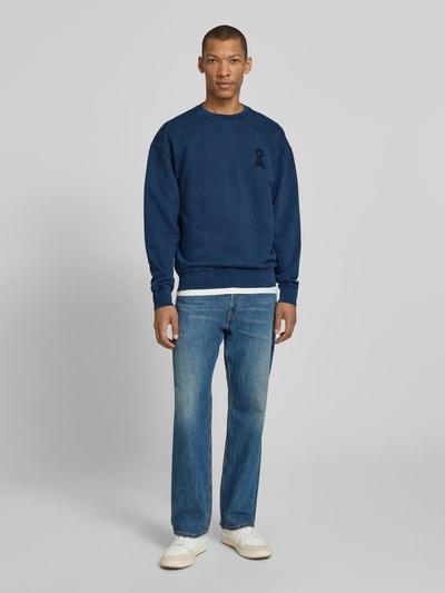 Męski model nosi Relaxed Fit Sweatshirt aus reiner Bio-Baumwolle Modell 'ICONIC Å GMT' od Armedangels, kolor granatowy, logo, długi rękaw, wygodny materiał, widok całej sylwetki z przodu.