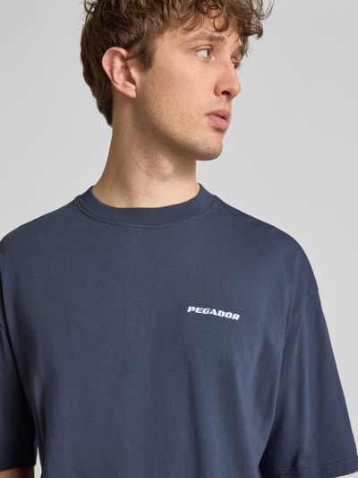 Männermodel trägt Oversized T-Shirt mit Logo-Stitching von Pegador aus Reine Baumwolle in Dunkelblau, Oversized-Fit, Rundhalsausschnitt, Nahaufnahme Oberkörper. Rückenlänge bei Größe M: 73 cm.