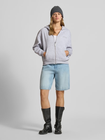 Frauenmodel trägt Sweatjacke mit Logo und Känguru-Tasche von REVIEW in Hellgrau, kombiniert mit Jeansshorts und schwarzen Stiefeln, Ganzkörperansicht von vorne. Baumwoll-Mix, Relaxed Fit, Reißverschluss, dezent melierte Textur.