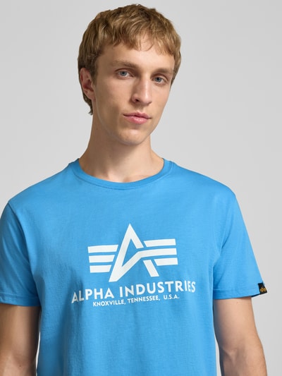 Mannenmodel draagt T-shirt met logo en ronde hals van Alpha Industries in effen lichtblauw, puur katoen, regular fit, ruglengte maat M 68 cm, Nahaufnahme Oberkörper.