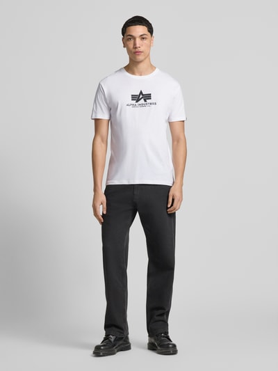 Männermodel trägt Herren T-Shirt aus Reiner Baumwolle Modell 'BASIC' von Alpha Industries in Weiß mit Label-Details, Gerader-Schnitt, Ganzkörperansicht von vorne.