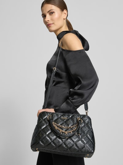 Frauenmodel trägt Handtasche in Leder-Optik Modell 'Libby' von Guess, Stepp-Optik, Logo-Detail, Zierbesatz, Magnetverschluss, Reißverschlusstasche, Tragehenkel, Abnehmbare Schulterriemen, Farbe Schwarz, glänzende Oberfläche, Größe: 32 cm Breite, 21 cm Länge, 11 cm Tiefe, Schulterriemen 122 cm, Ganzkörperansicht von vorne.