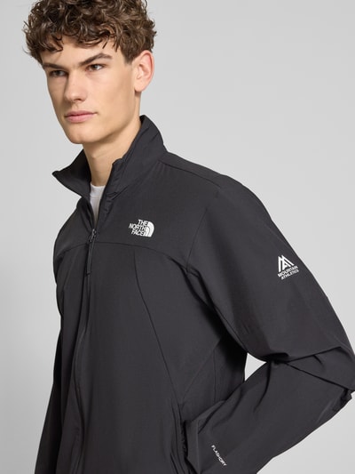 Męski model nosi Regular Fit Jacke mit Stehkragen Modell 'Mountain' von The North Face, czarny, elastyczna mieszanka materiałów, nadruk z logo, zamek błyskawiczny, wpuszczane kieszenie, elastyczne wykończenia, długość tyłu w rozmiarze M 73 cm, szerokość ramion w rozmiarze M 43 cm, szerokość klatki piersiowej w rozmiarze M 56 cm, długość rękawów w rozmiarze M 70 cm, półzbliżenie na górną część ciała z przodu