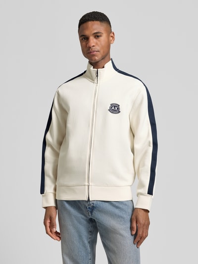 Męski model nosi Sweatjacke mit Zweiwege-Reißverschluss Modell 'FELPA' od ARMANI EXCHANGE z bawełnianej mieszanki, krój regular fit, kremowa z granatowymi detalami, label-stitching, stojący kołnierz, długość tyłu w rozmiarze M: 67 cm, długość rękawa w rozmiarze M: 61 cm, widok całej sylwetki z przodu.