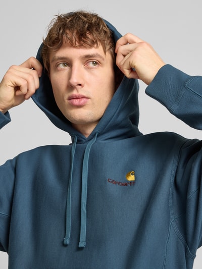 Mężczyznamodel nosi Hoodie mit Kapuze von Carhartt Work In Progress w kolorze niebieskim, mieszanka bawełny, luźny krój, kaptur, tunel, wyhaftowane logo, kieszeń kangurka, prążkowane wykończenia, długość tyłu 70 cm, długość rękawa 64 cm, zbliżenie na górną część ciała.