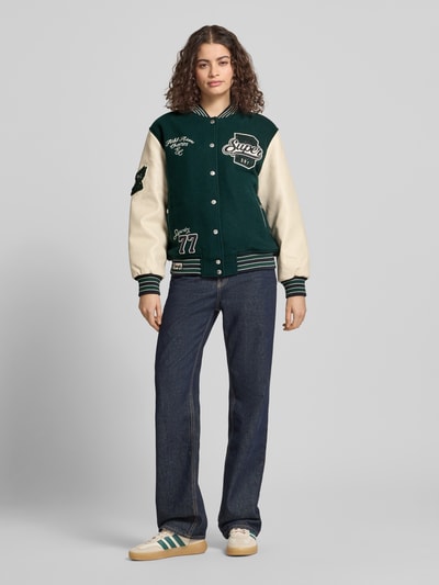 Frauenmodel trägt College Jacke mit Motiv- und Logo-Stitchings von Superdry im Woll-Mix, Dunkelgrün-Beige, Lockerer-Schnitt, Stehkragen, Druckknopfverschluss, elastische Abschlüsse, Rückenlänge bei Größe S 62 cm, Schulterbreite 49 cm, Brustweite 60 cm, Ärmellänge 60 cm, Ganzkörperansicht von vorne.