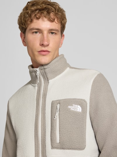 Męski model nosi kurtkę z polaru z wysoką stójką Model 'YUMIORI' od The North Face, jasnoszary i kremowy kolor, kieszeń na zamek błyskawiczny na piersi, luźny krój, komfort noszenia, Długość tyłu w rozmiarze M: 71 cm, Długość rękawów w rozmiarze M: 71 cm, zbliżenie na górną część ciała.