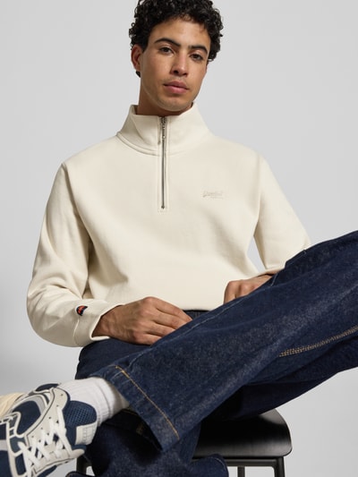 Männermodel trägt Regular Fit Sweatshirt mit Logo-Stitching von Superdry, cremefarbener Baumwoll-Mix, Stehkragen mit Reißverschluss, elastische Abschlüsse, Ganzkörperansicht von vorne.