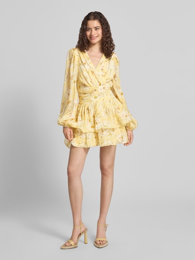 Frauenmodel trägt Minikleid aus Viskose-Mix mit Rüschenbesatz von Gina Tricot in Gelb mit floralem Muster, taillierte Passform, V-Ausschnitt, langärmlig, Ganzkörperansicht von vorne.